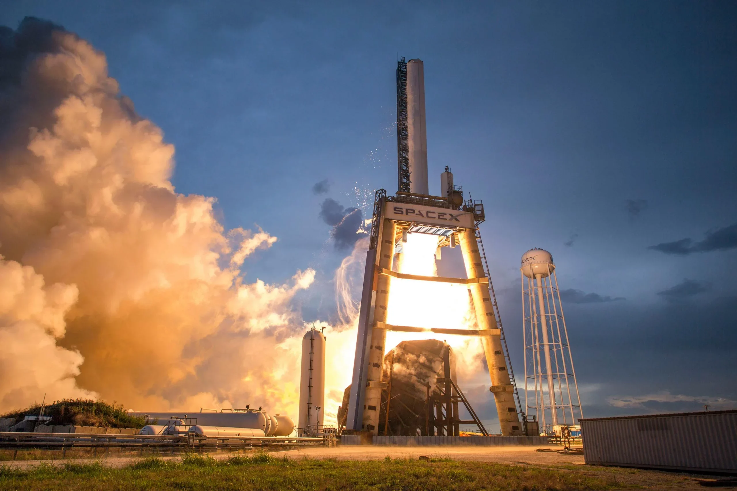 Rocket test stand with a bright flame and a large orange smoke cloud at dusk, SpaceX lettering on the tower