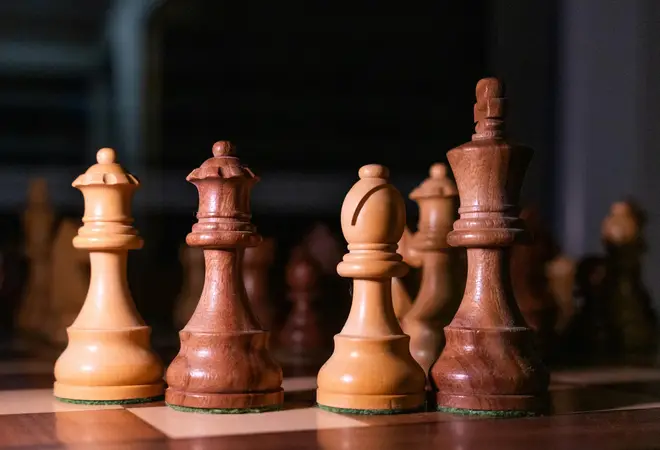 Wooden chess pieces including a king, queen and bishop on a board with more pieces blurred behind