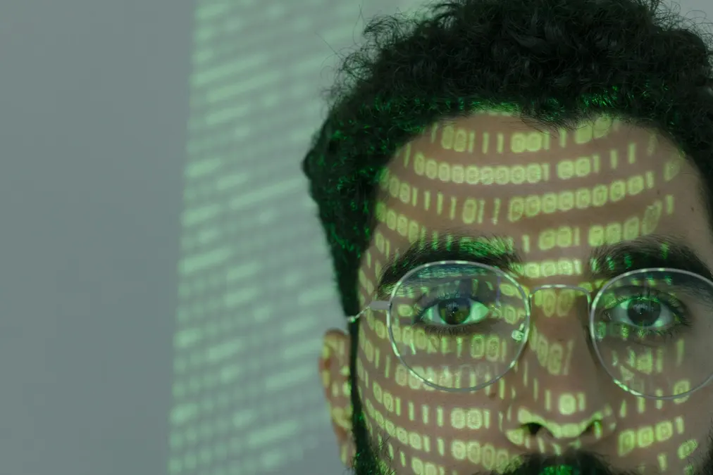 Close-up of a man's face with glasses lit by projected green binary code