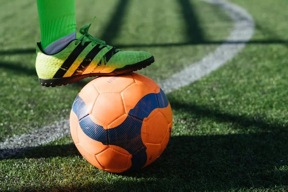 Foot in a green cleat resting on an orange and blue football on artificial turf