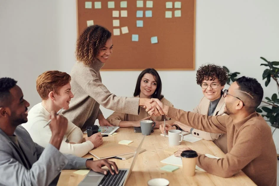 Smiling colleagues shaking hands across a wooden table in front of a board with sticky notes