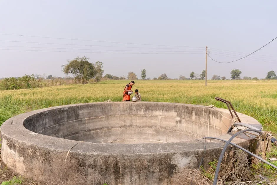 Woman in orange sari with a child filling a pot beside a large empty round concrete well in a green field