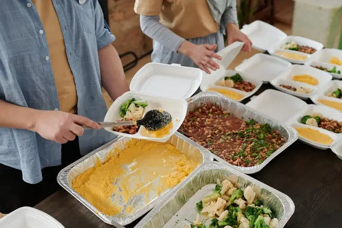 Volunteers serving cooked vegetables and lentils from foil trays into takeaway food boxes on a table