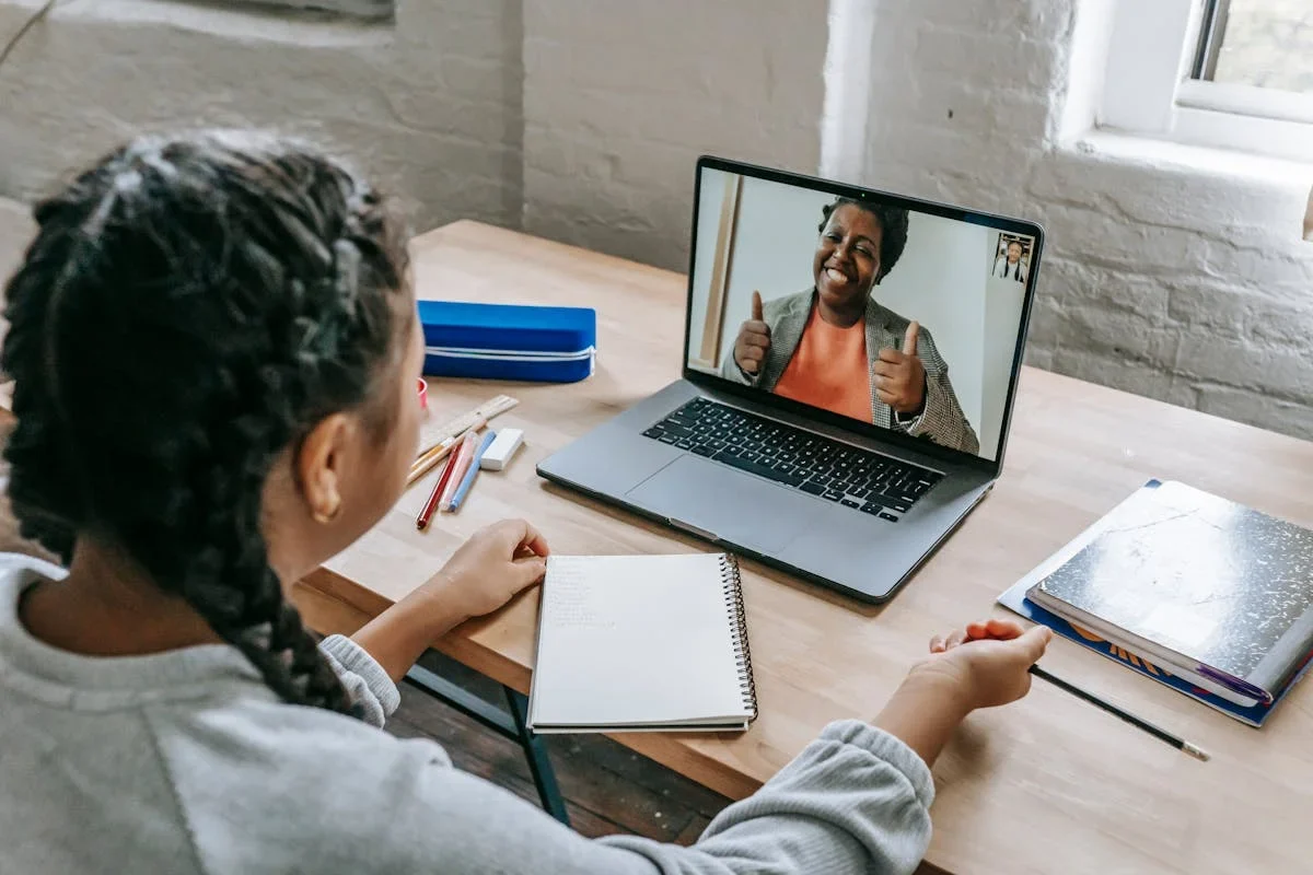 Girl with braided hair at a wooden desk on a video call with a smiling woman giving thumbs up on a laptop