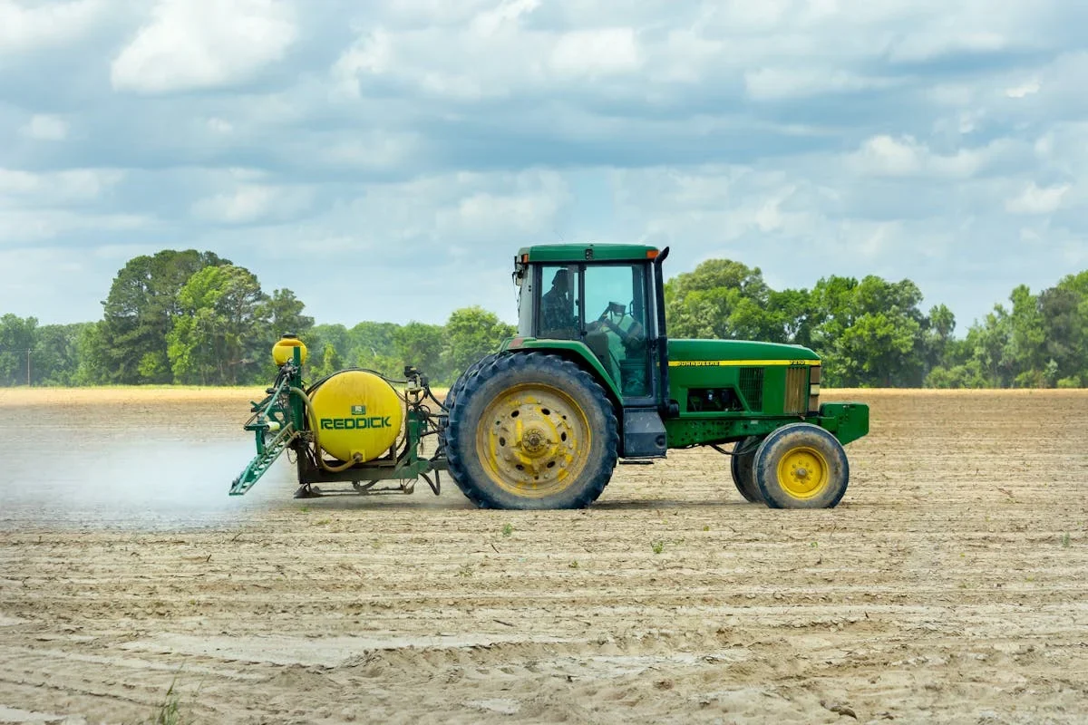 Green tractor with a yellow sprayer tank crossing a dry field under a cloudy sky