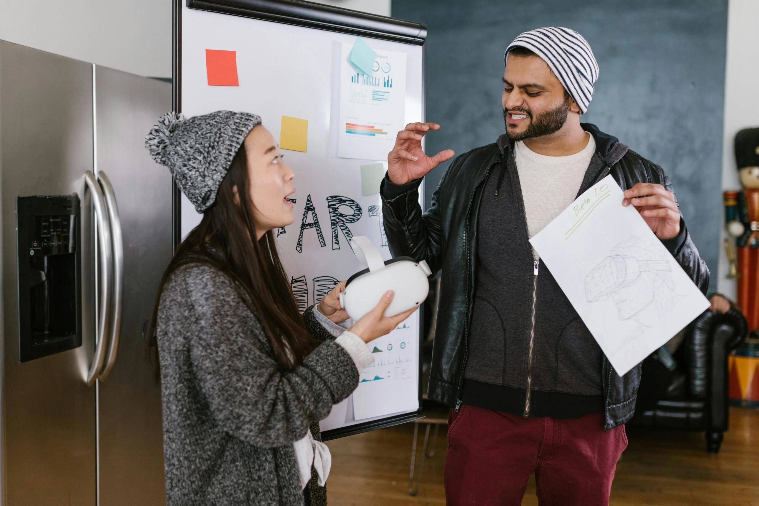 Two young people in beanies talking, one holding a VR headset and the other a sketch, in front of a whiteboard