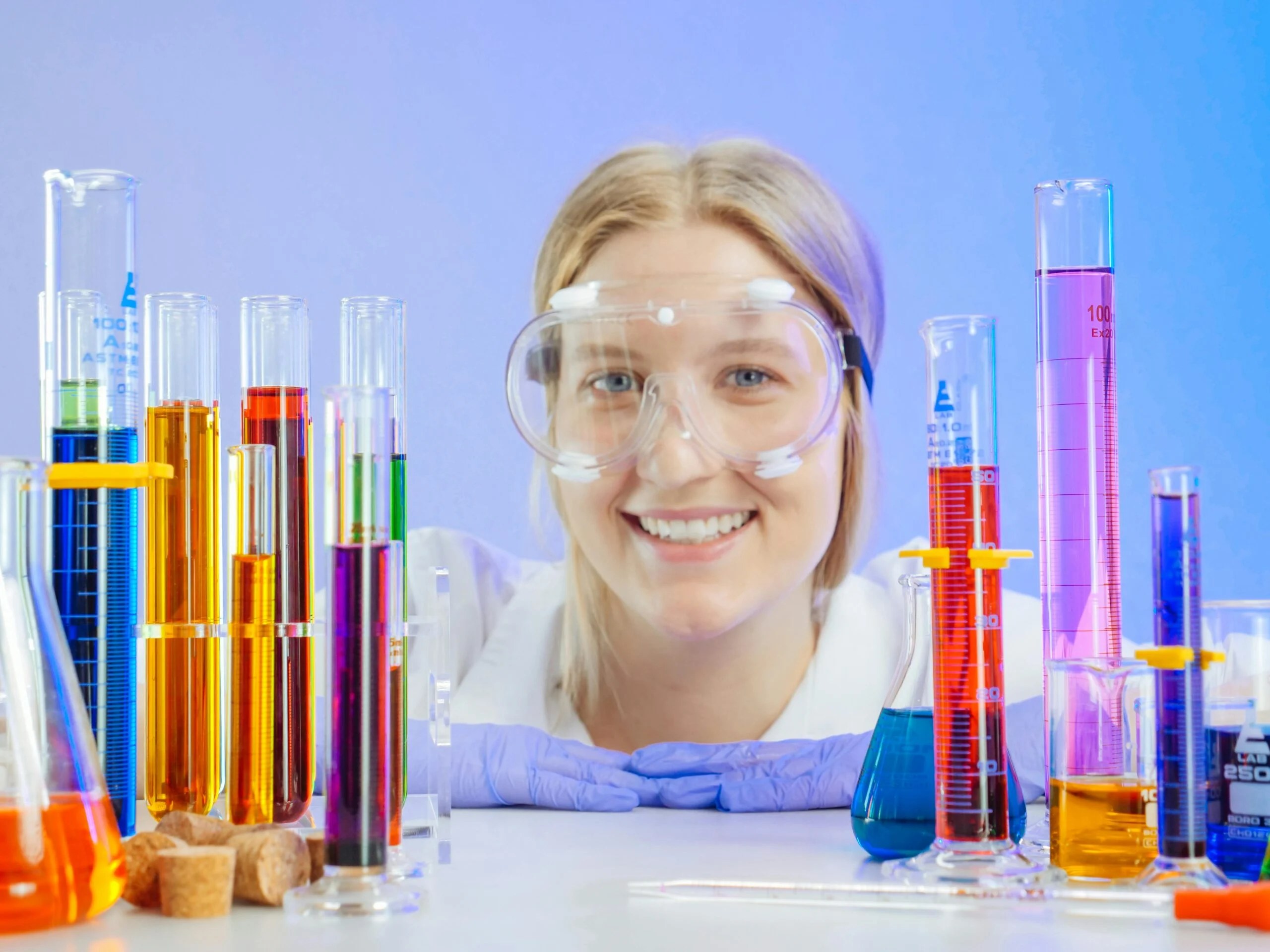 Smiling woman in safety goggles and lab coat behind colourful liquids in glass measuring cylinders