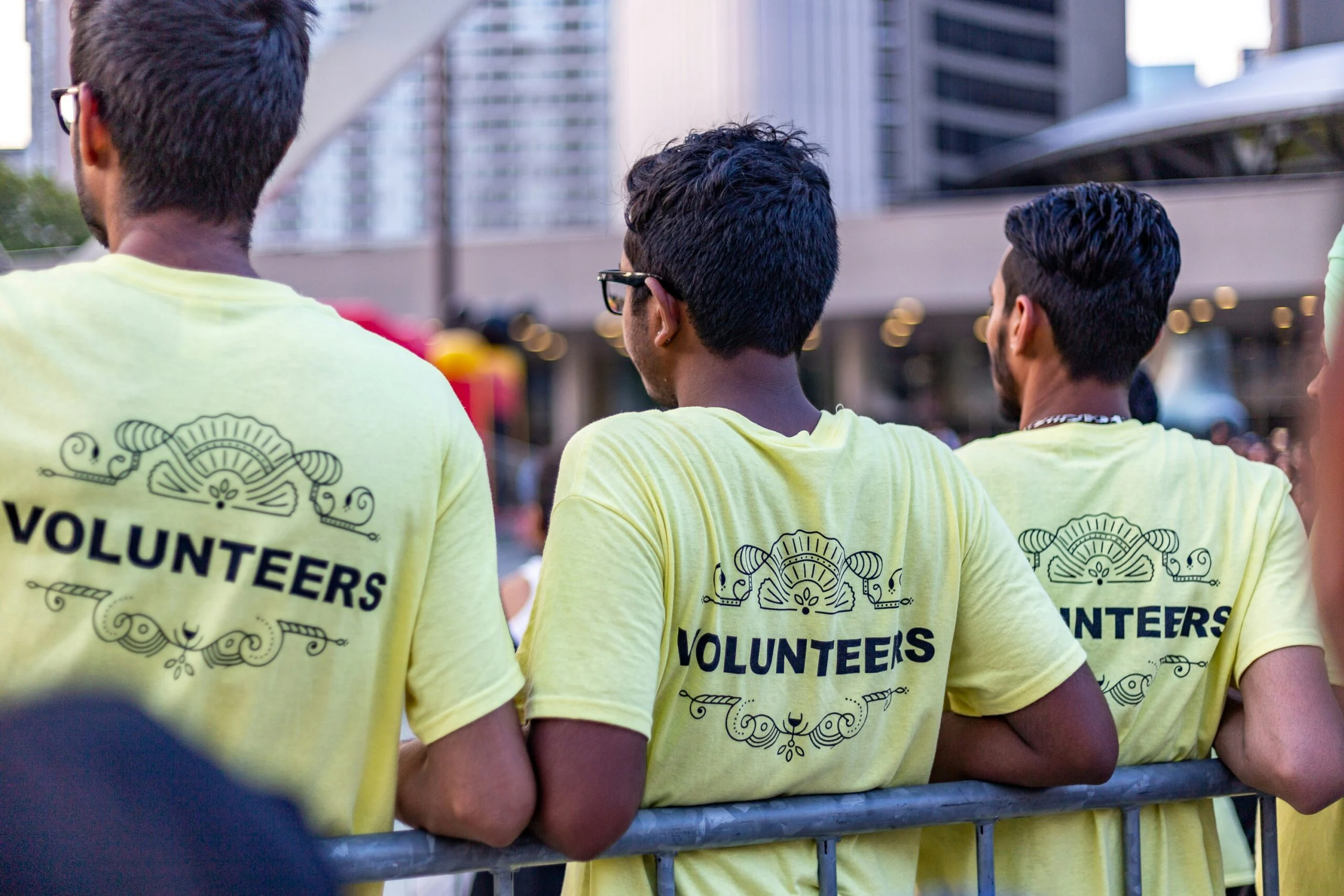 Three young men seen from behind in yellow Volunteers t-shirts leaning on a metal barrier in a city plaza