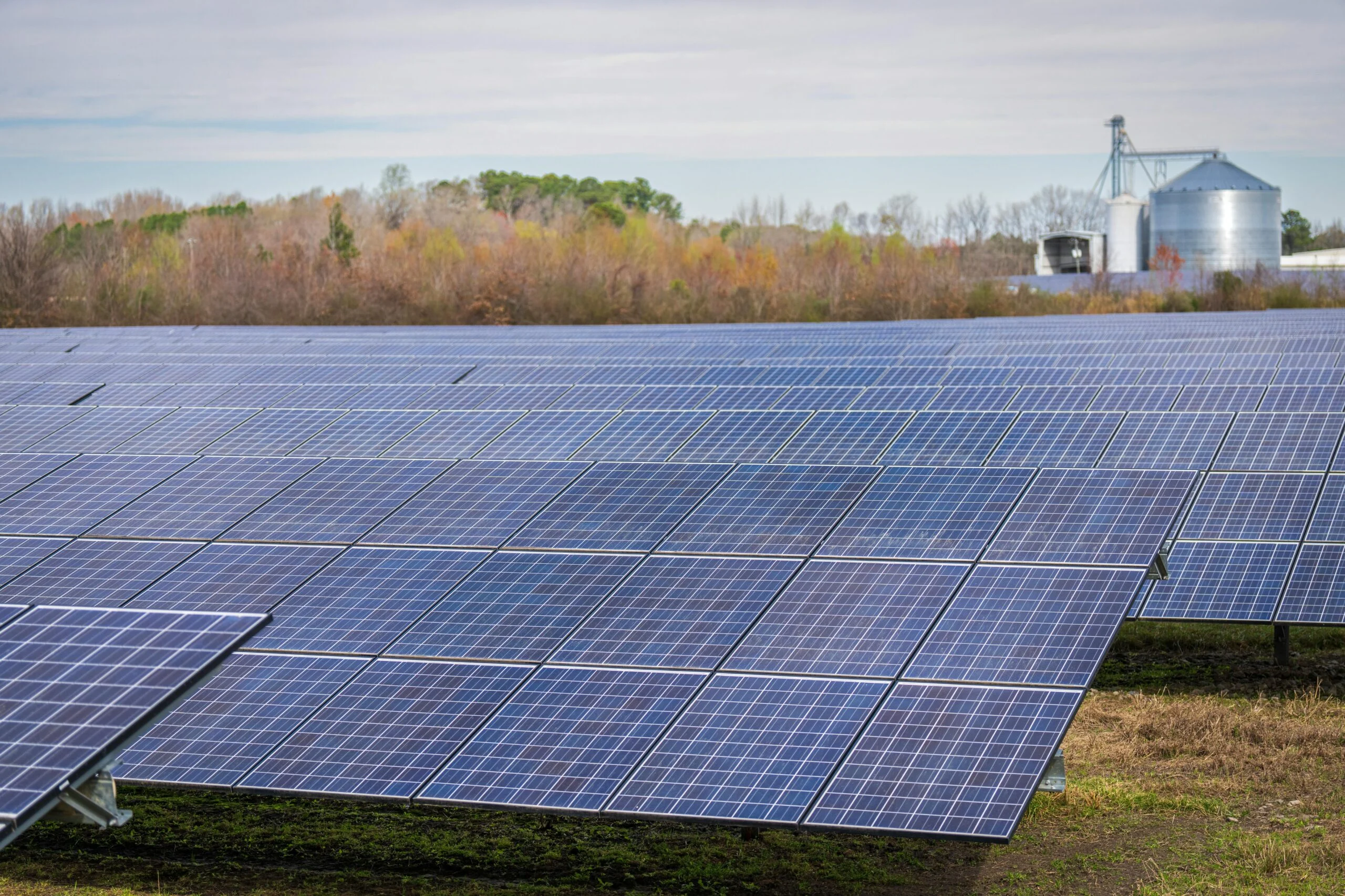 Rows of solar panels in an open field with trees and a grain silo in the background