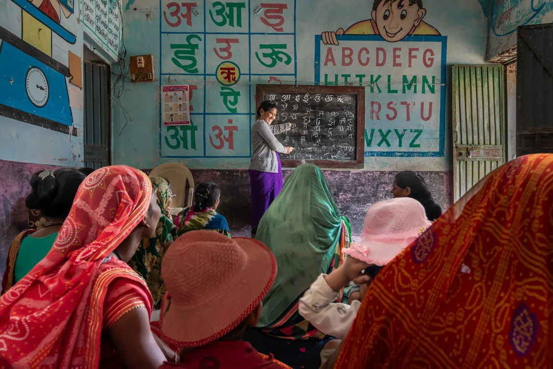 Woman teacher writing on a blackboard in a painted classroom while women in colourful saris watch