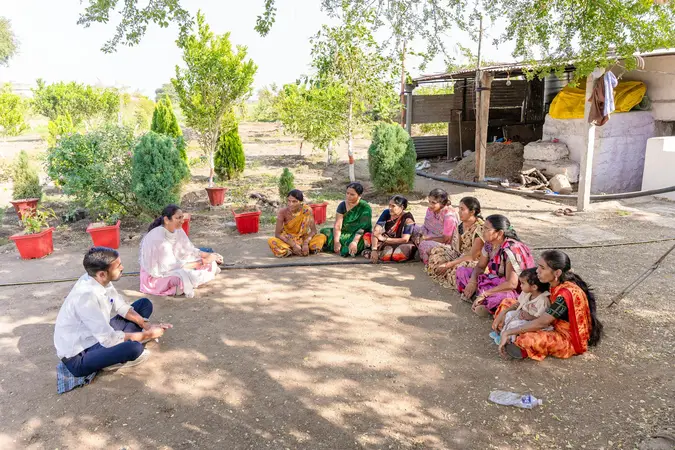 Women sitting in a circle on the ground in a village yard with a man and a woman facing them, plants in red pots behind