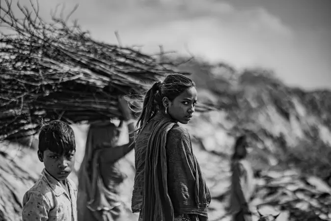 Black and white photo of a girl looking over her shoulder and a boy, with a person carrying branches behind