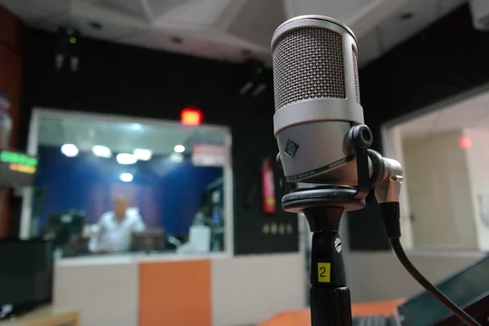 Studio microphone in the foreground with a presenter visible through the glass of a radio booth behind