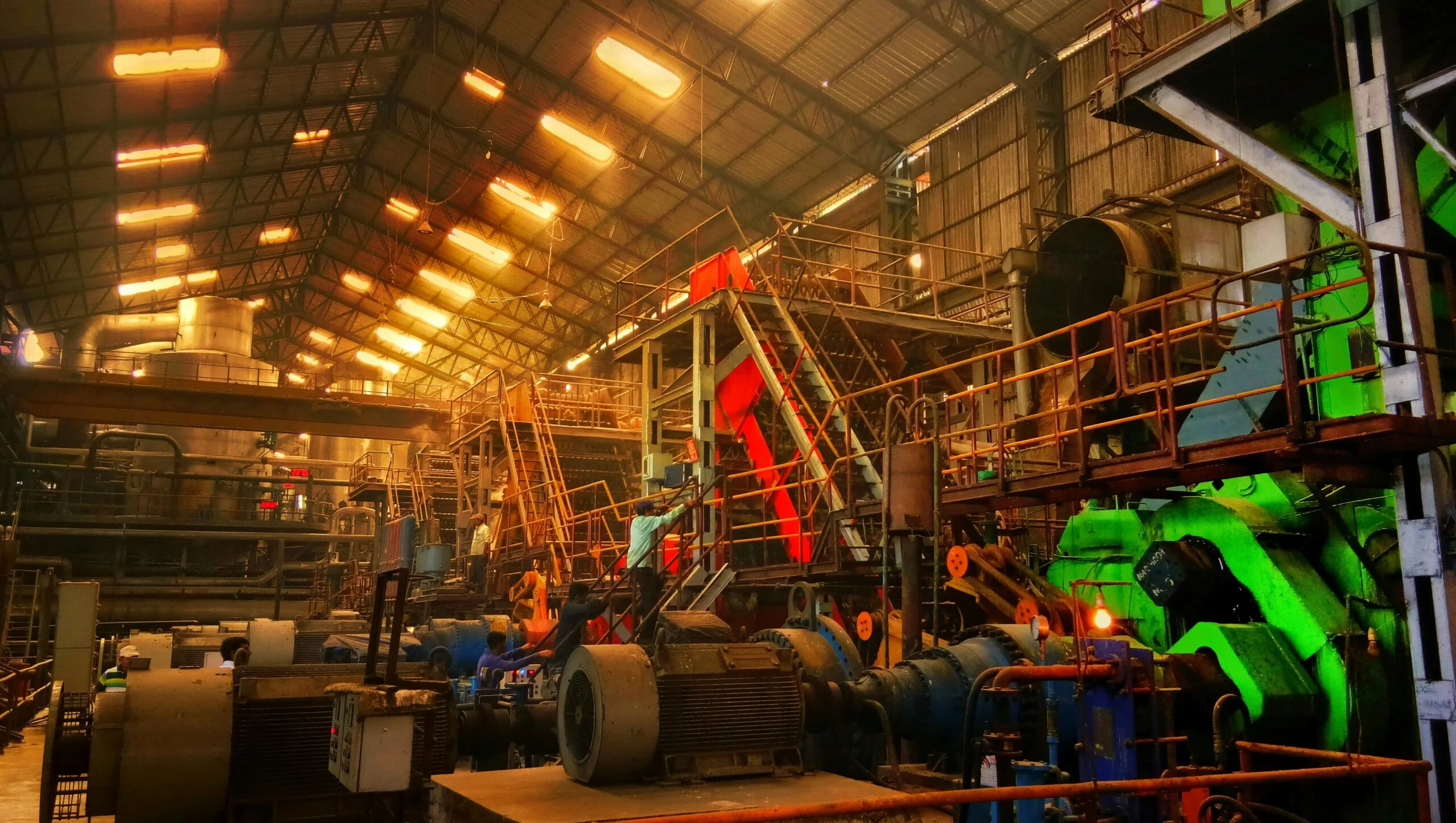 Inside a steel plant with overhead lights, orange and green machinery, gantries and workers on the factory floor