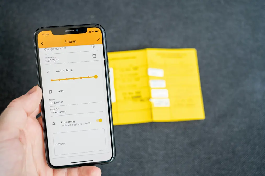 Hand holding a smartphone showing a vaccination record app beside a yellow paper vaccination booklet