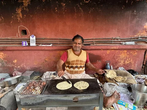 Smiling street vendor in a patterned vest cooking flatbreads on a griddle by a red wall with a QR code sticker
