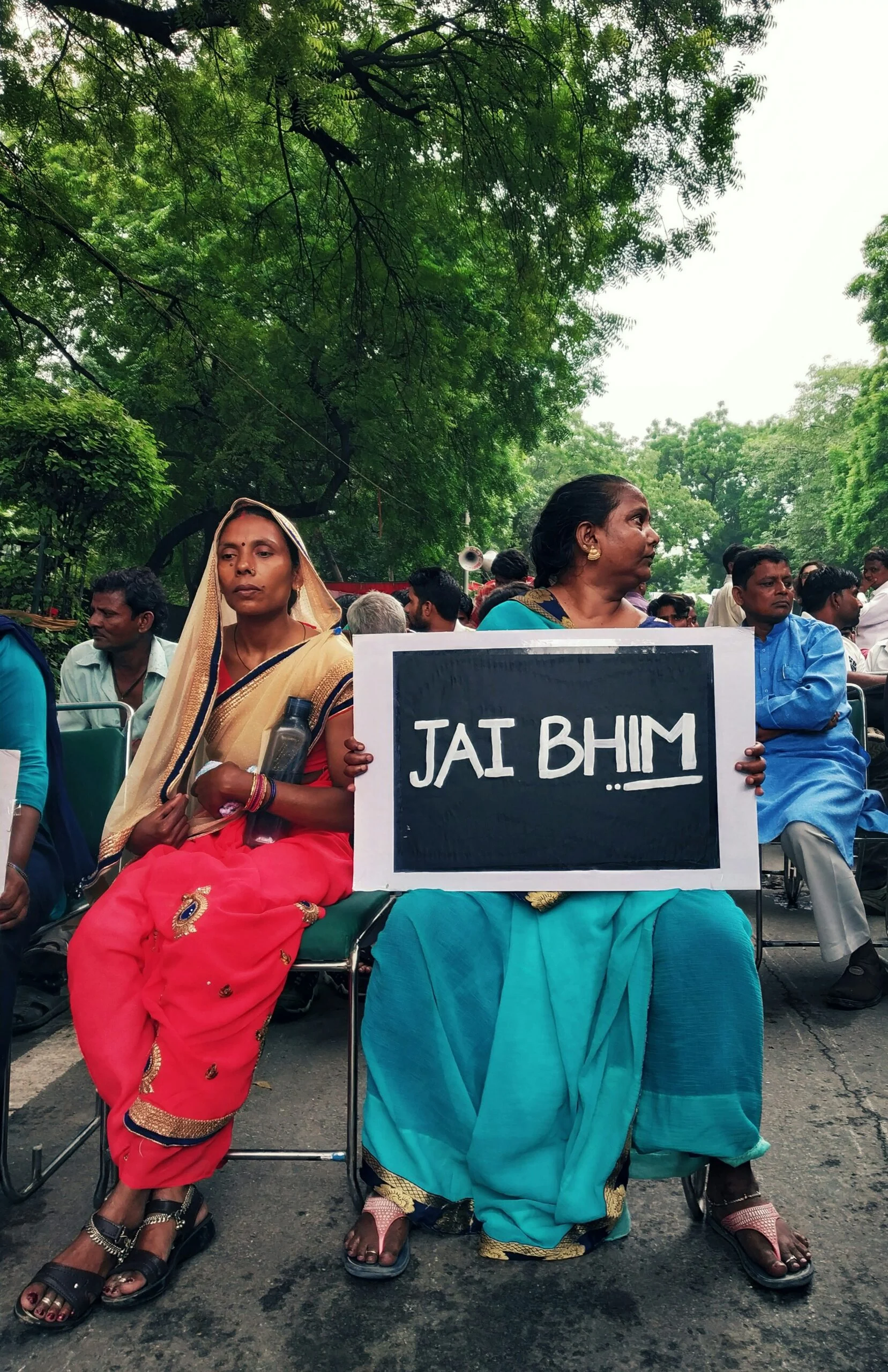 Two women in saris seated outdoors at a protest, one holding a black sign reading JAI BHIM, with trees behind