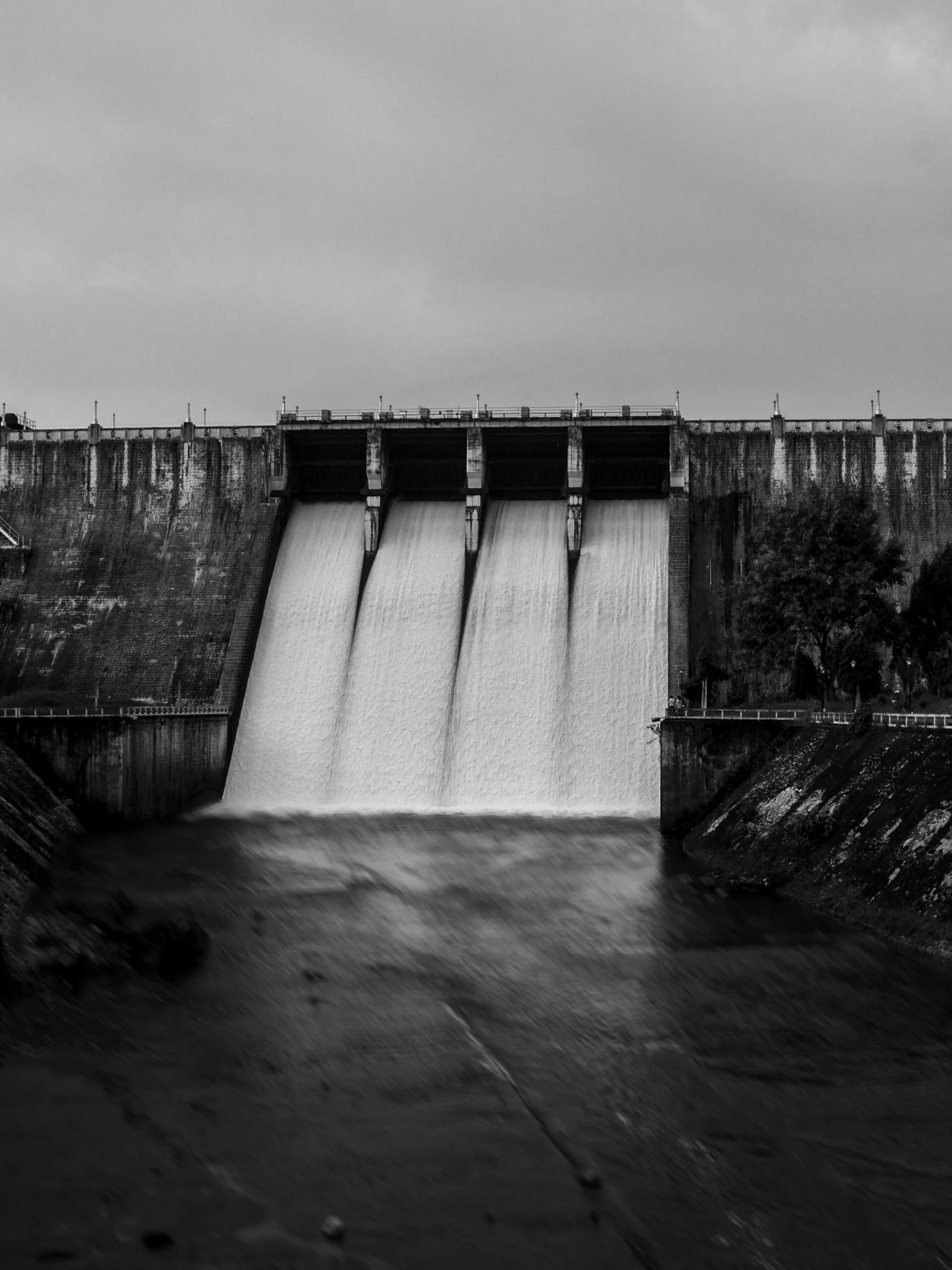 Black and white photo of a concrete dam with water spilling through four open spillway gates