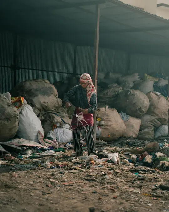 Waste picker in a headscarf standing among large sacks of sorted rubbish under a corrugated metal shelter