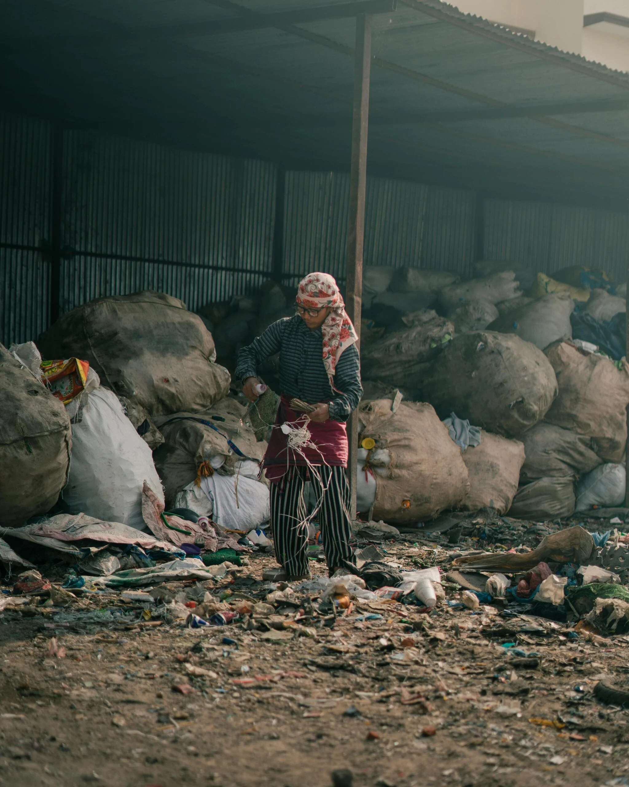 Waste picker in a headscarf standing among large sacks of sorted rubbish under a corrugated metal shelter