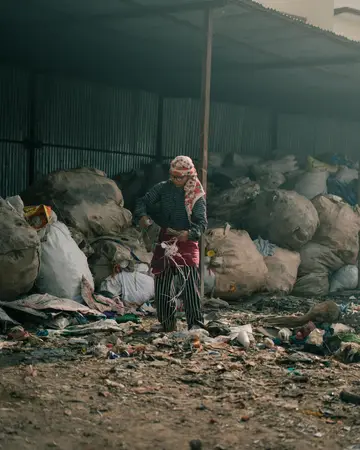 Waste picker in a headscarf standing among large sacks of sorted rubbish under a corrugated metal shelter