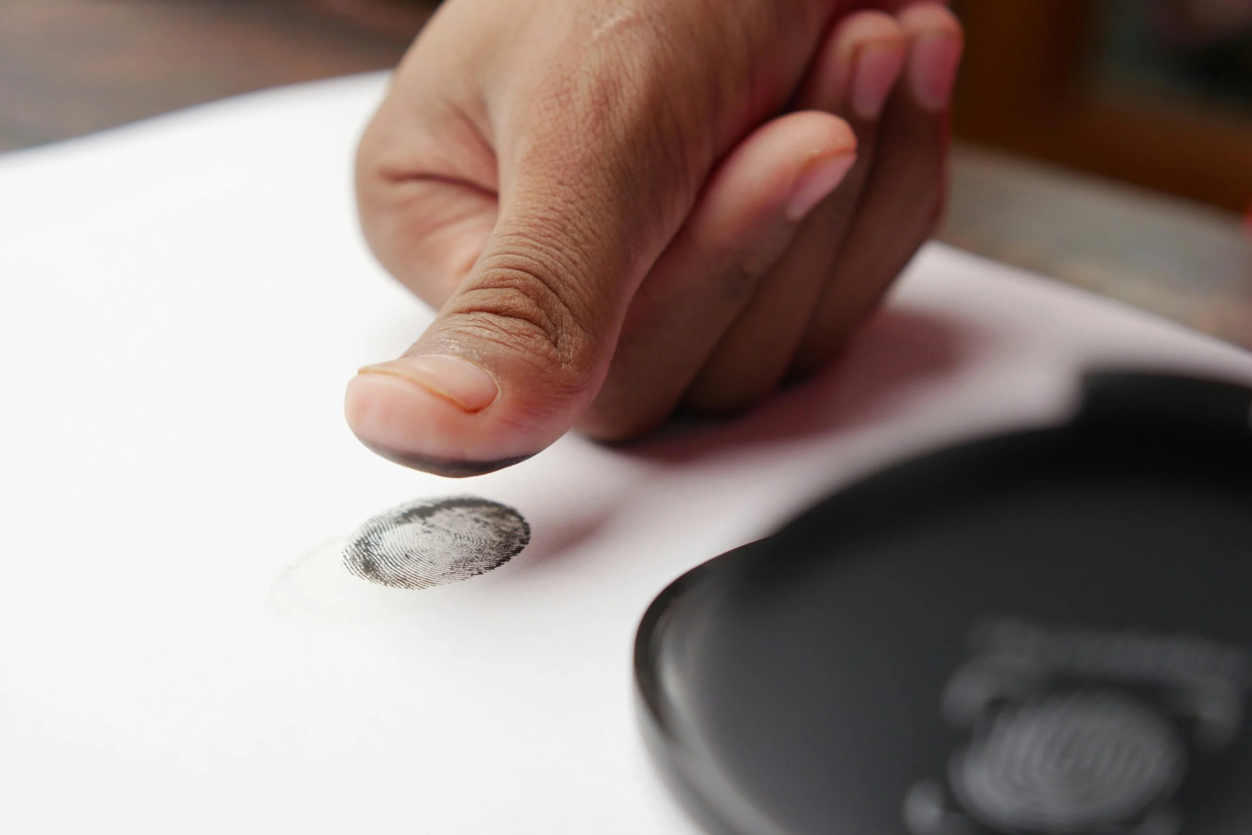 Inked thumb pressed onto white paper leaving a fingerprint, with an ink pad beside it