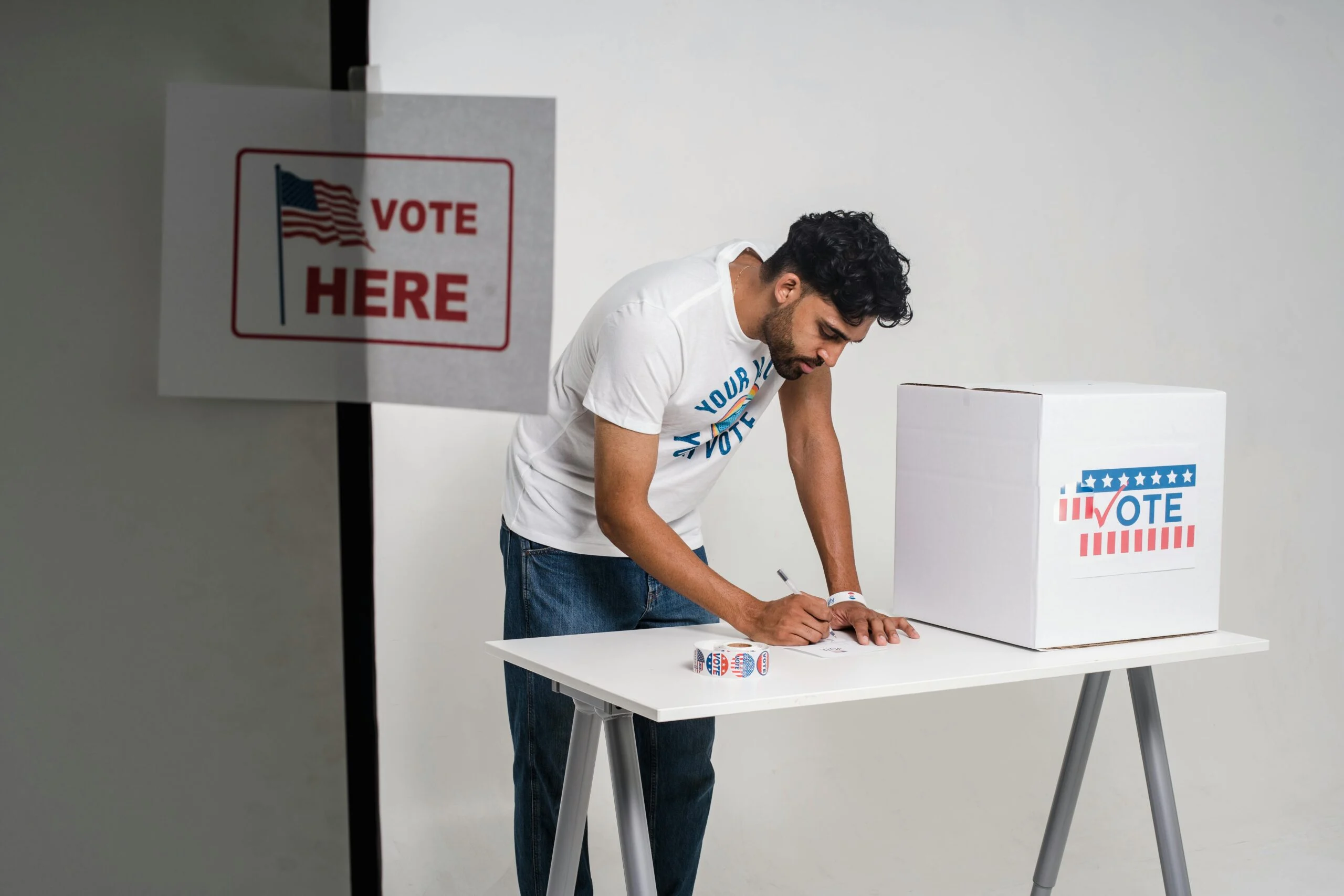 Man writing at a white table beside a ballot box marked Vote, with a Vote Here sign showing a US flag