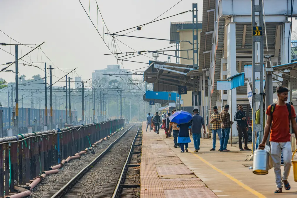Railway platform with passengers walking, one under a blue umbrella, beside overhead wires and a track in morning haze