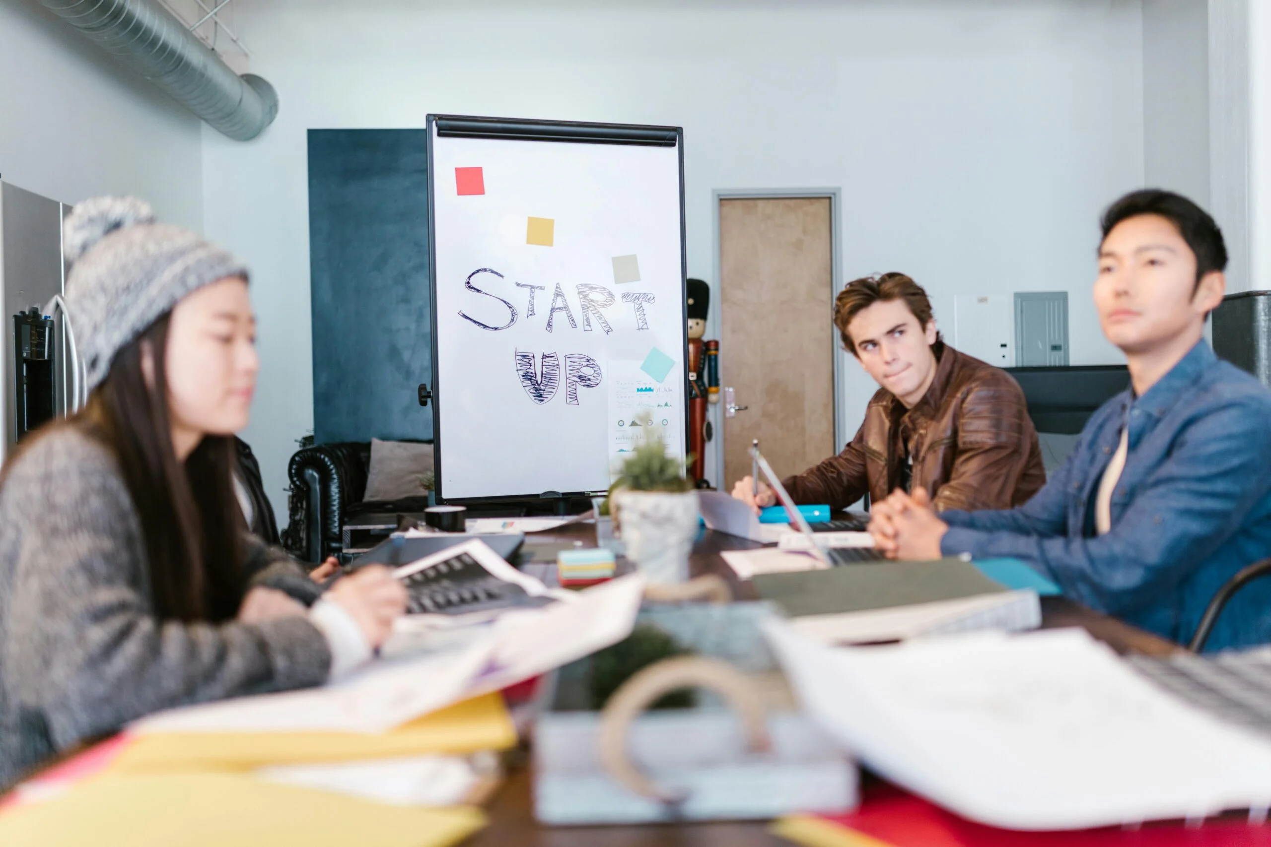 Three people at a meeting table with papers and laptops in front of a whiteboard reading Startup
