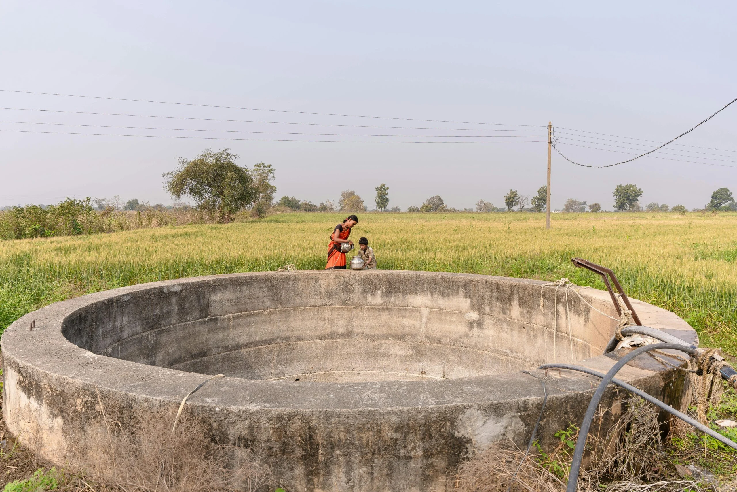Woman in orange sari and a child filling a pot beside a large empty round concrete well in a green field
