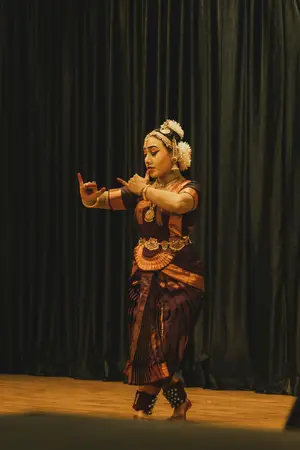 Bharatanatyam dancer in a maroon and gold costume striking a hand gesture on a stage before a dark curtain