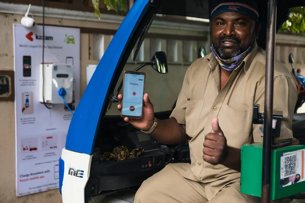 Smiling auto-rickshaw driver in khaki uniform holding a phone showing Charging, next to an EV charging point on a wall