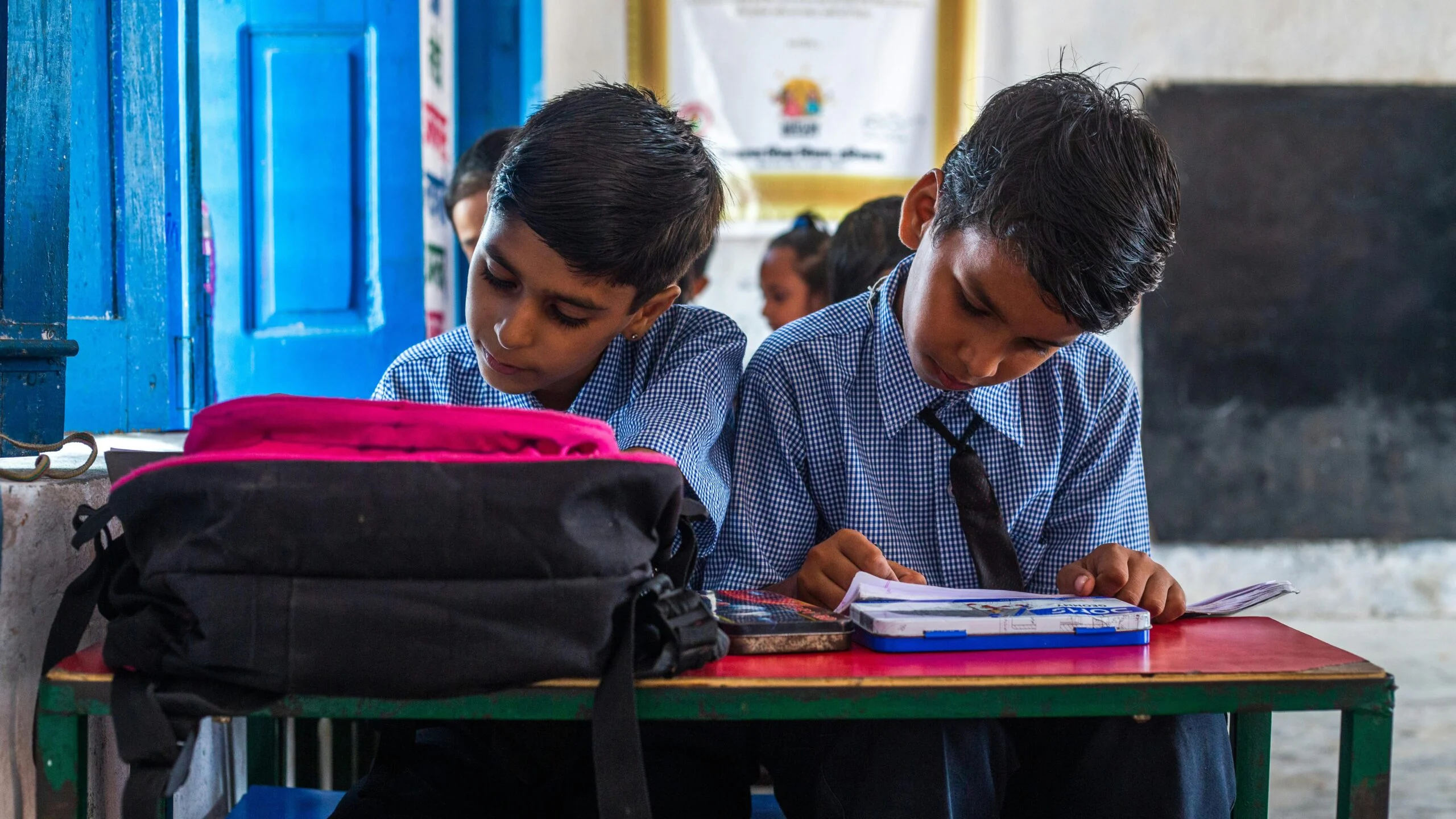 Two schoolboys in checked uniforms and ties reading an open book at a red desk with a black backpack in front of them
