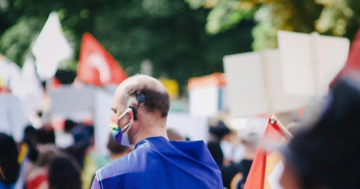 Man in a blue jacket wearing a rainbow face mask seen from behind at a march with flags and blank placards