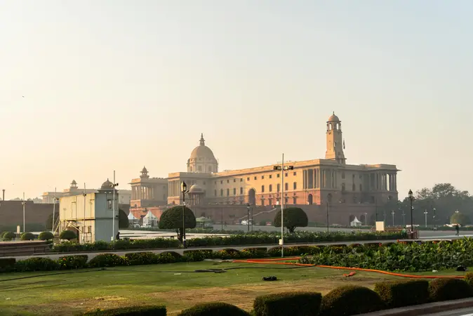 Sandstone government building with domes and a clock tower seen across lawns and hedges in morning haze