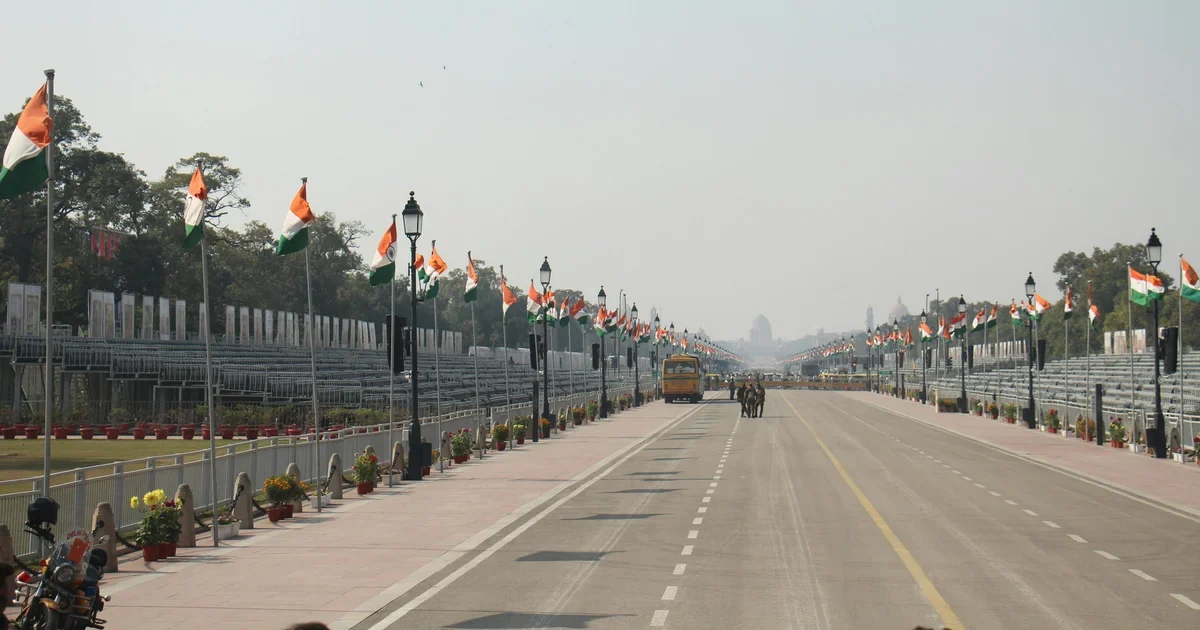 Wide empty road lined with saffron, white and green flags and spectator stands, with a bus in the distance