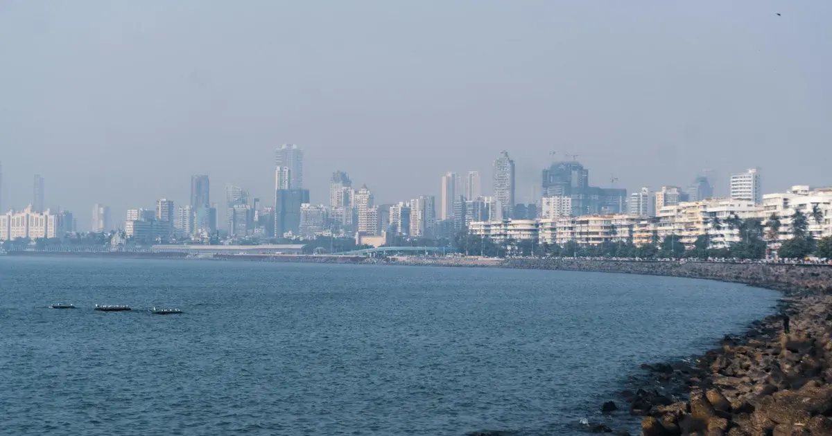 Mumbai skyline of tall apartment towers seen across the sea from a rocky shoreline on a hazy day