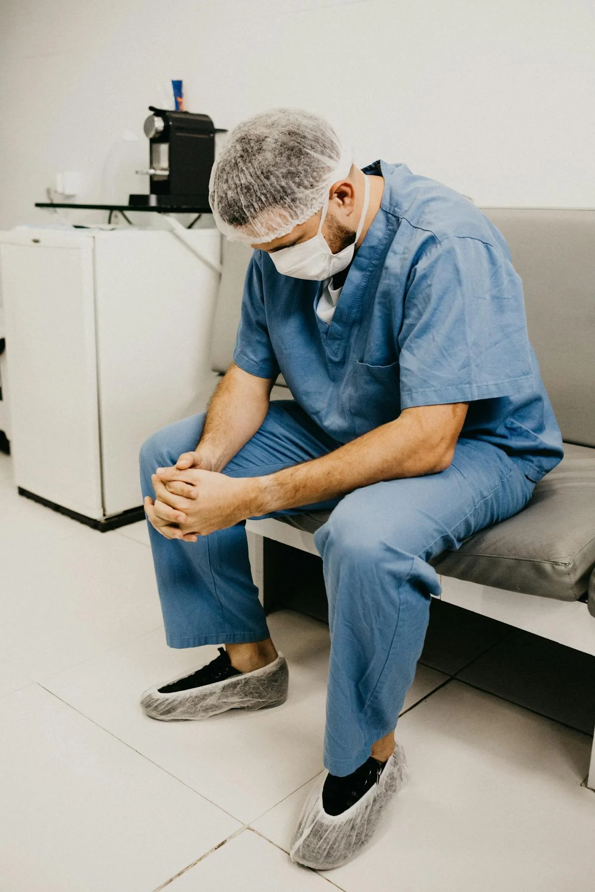 Surgeon in blue scrubs, mask and shoe covers sitting on a bench with head bowed and hands clasped