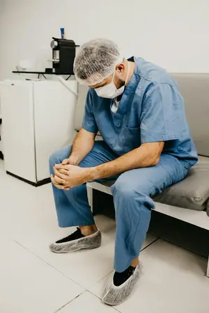 Surgeon in blue scrubs, mask and shoe covers sitting on a bench with head bowed and hands clasped
