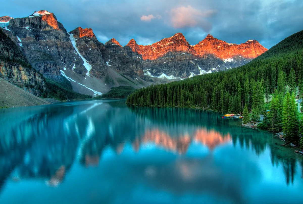 Alpine lake with a mountain reflected in turquoise water, rocky shoreline, and orange-lit peaks beyond pine forest
