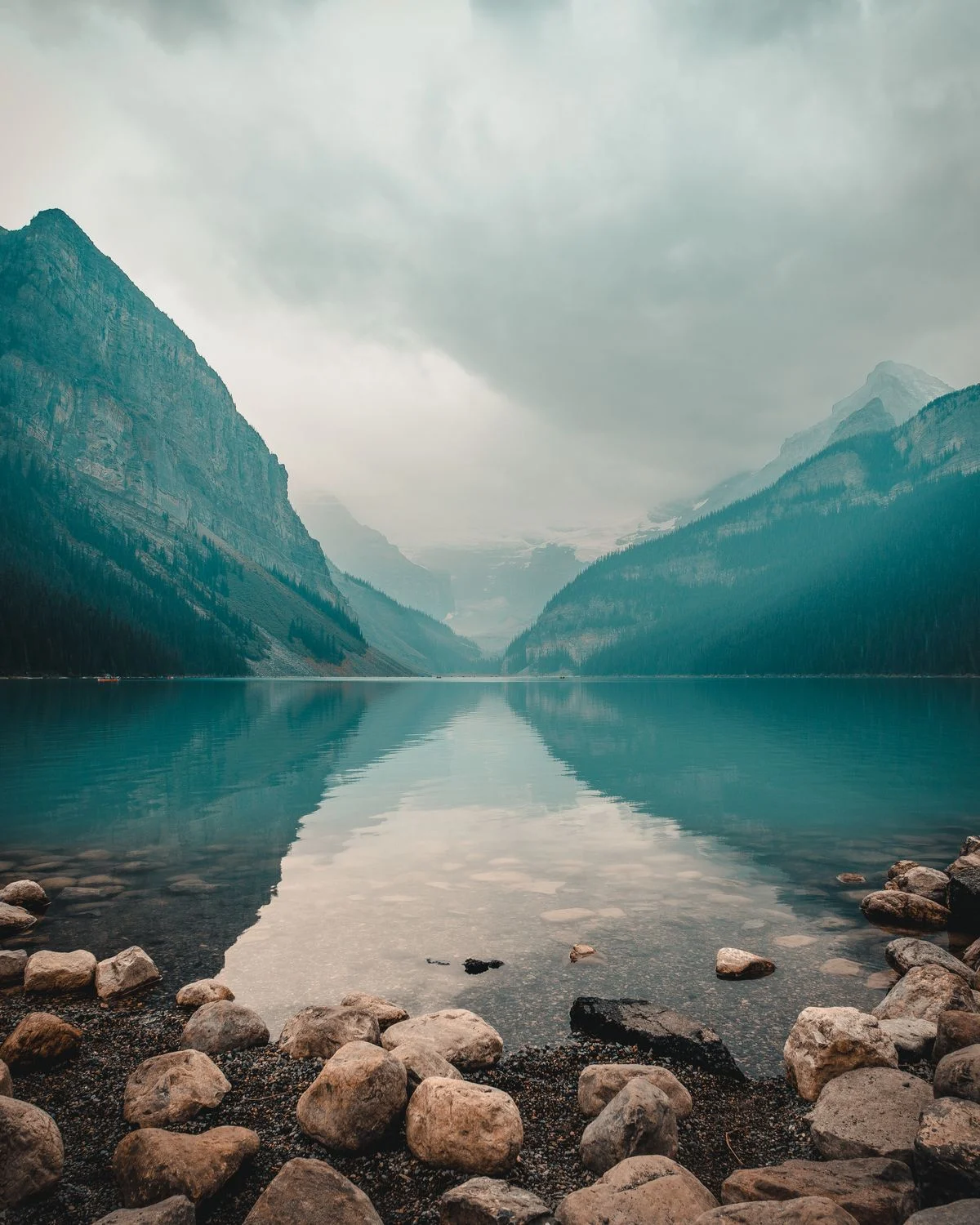 Misty mountain lake with steep forested slopes reflected in calm green water and smooth stones along the shore