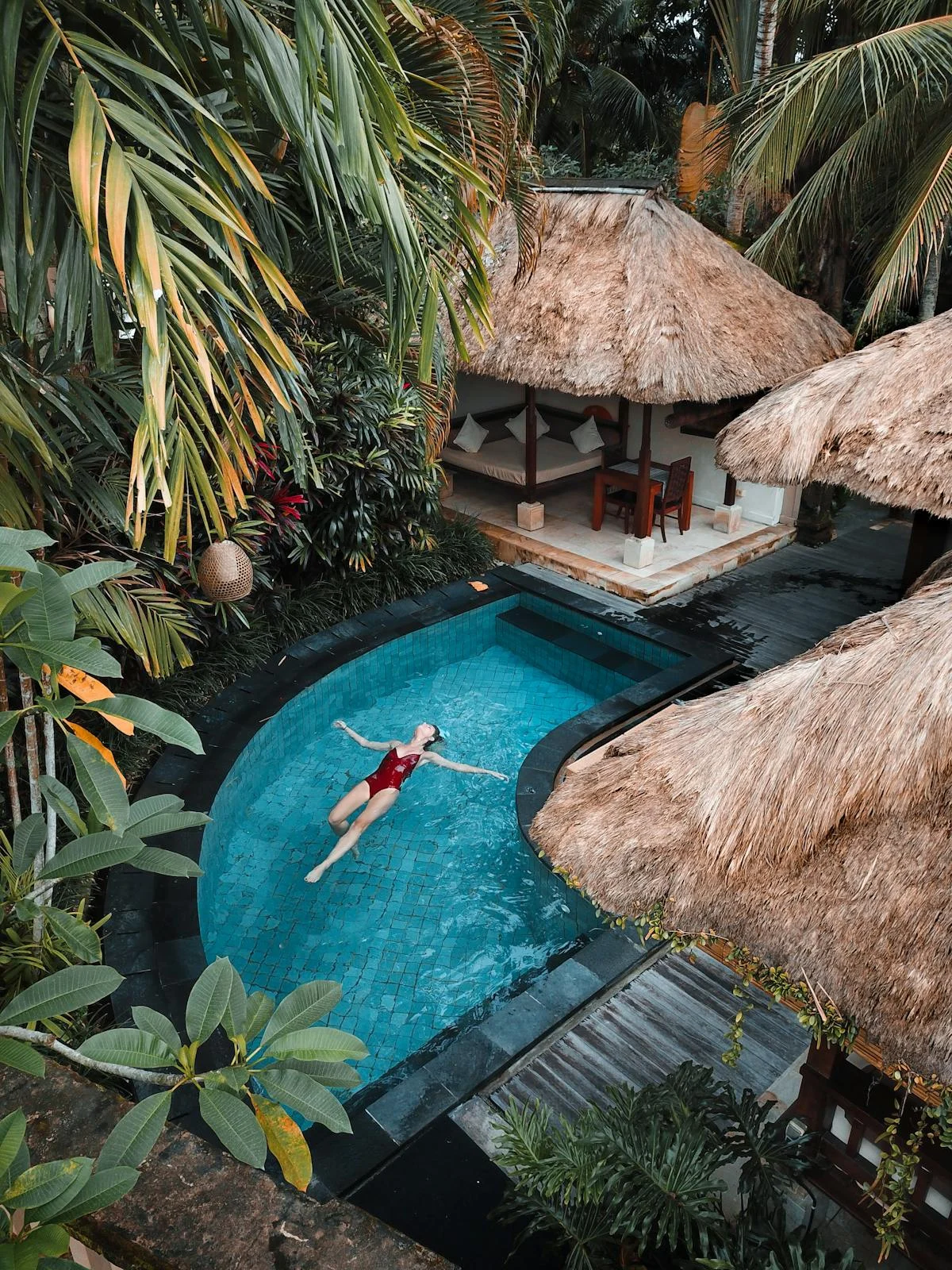 Aerial view of a curved turquoise swimming pool with a woman floating, thatched huts and palm trees around it