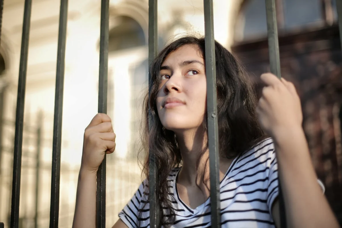Young woman in a striped t-shirt holding the bars of a metal fence and looking upward