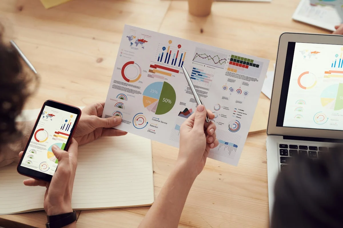 Hands holding a printed sheet of pie charts and bar graphs beside a phone and laptop showing the same charts
