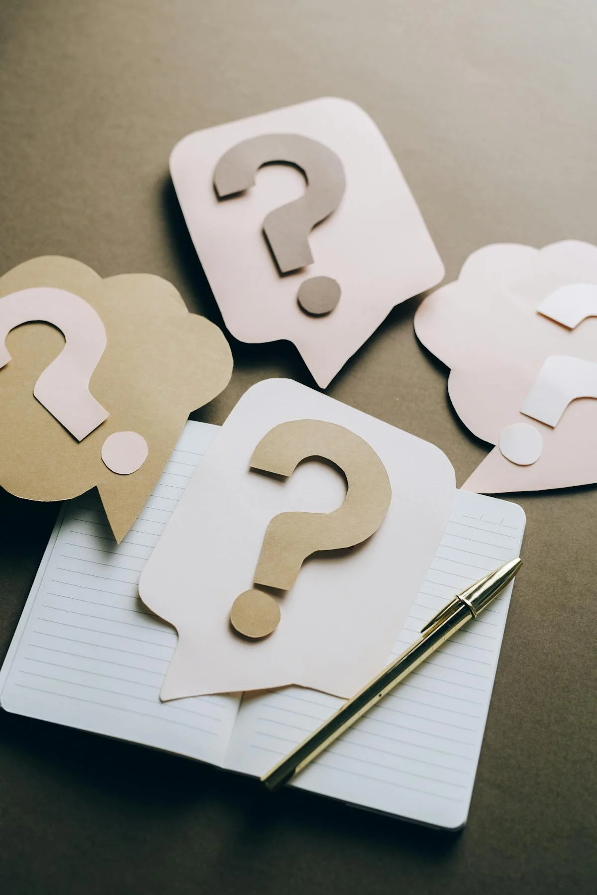 Paper cutout speech bubbles with question marks on an open lined notebook beside a gold pen