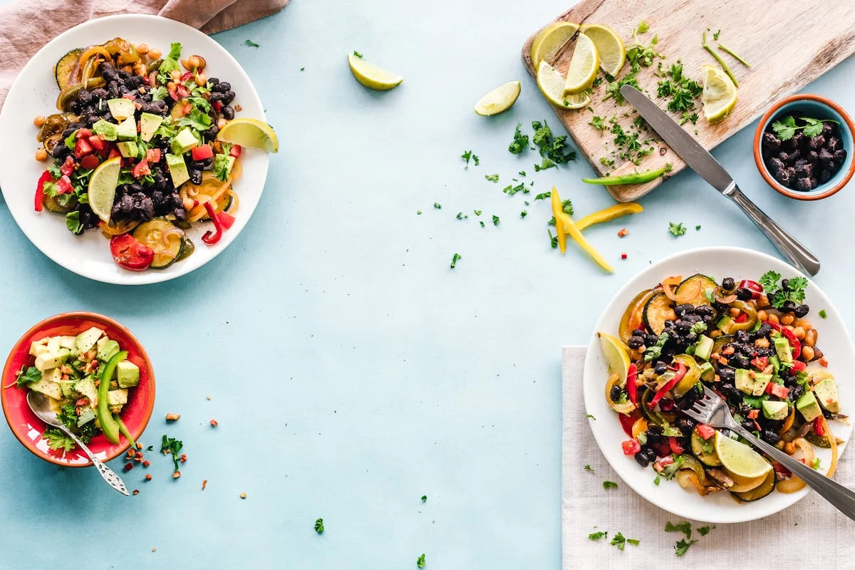 Two white plates and a bowl of black bean, avocado and vegetable salad with lime and herbs on a light blue table