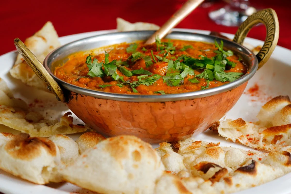 Copper bowl of orange curry garnished with coriander, surrounded by pieces of toasted flatbread on a white plate