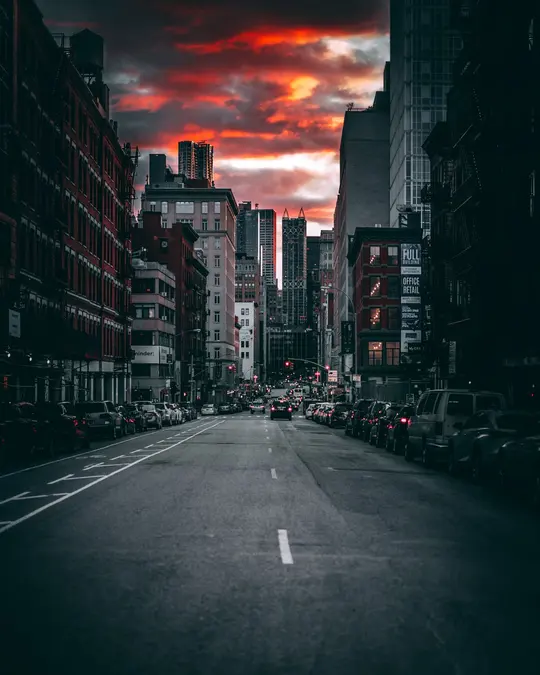 City street between tall brick buildings at sunset with red clouds and parked cars along both kerbs