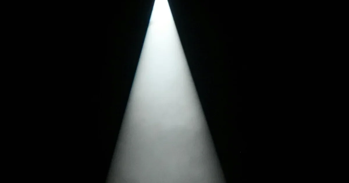 Cone of white spotlight shining down through darkness in an empty black space