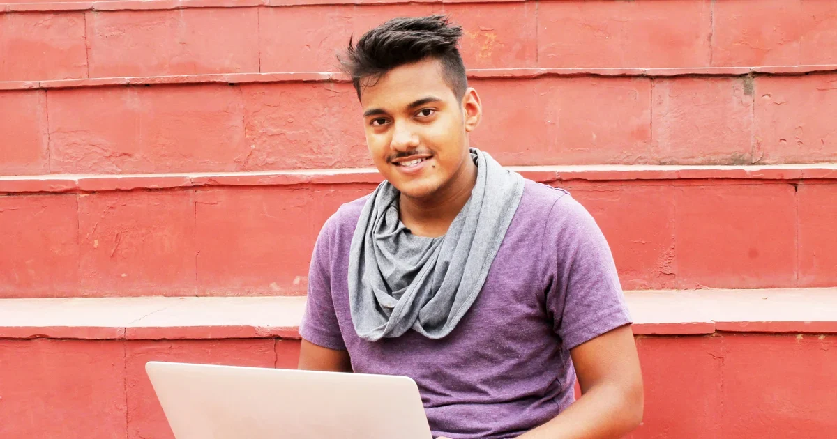 Smiling young man in a purple t-shirt and grey scarf sitting on red steps with a laptop